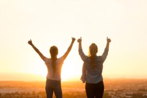 silhouette of two women with hands in the air in celebration facing a sunset 