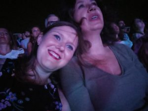 A woman smiling leaning her head on a woman having an emotional moment in a huge crowd at a live concert