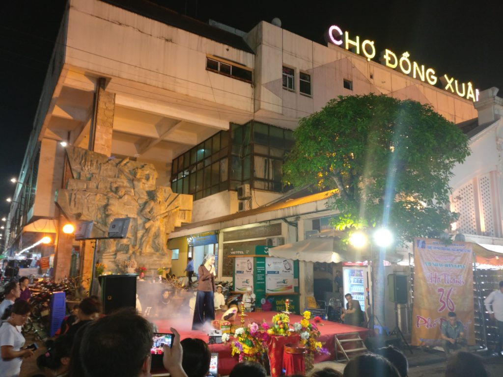 A woman is on stage at the Dong Xuan Night Market singing. She is wearing plain black pants and a brown top. The view is not great as there are many distractions around.