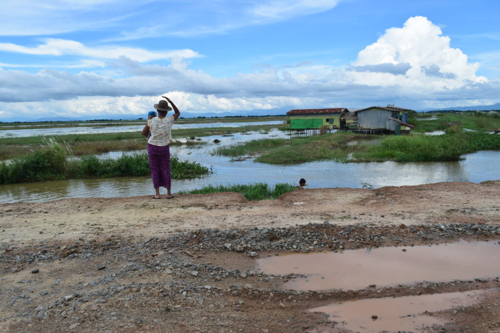 a view of a rough road in Myanmar with a woman holding a baby looking out over rice paddy